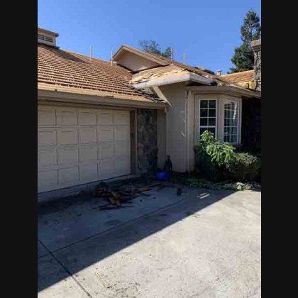 House with damaged roof and garage door; debris on the driveway. Sunny day.