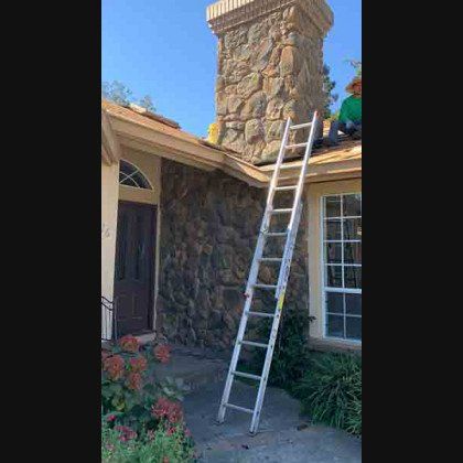 Ladder leaning against a house with a stone chimney; a worker is on the roof.