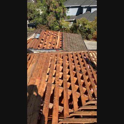 Rooftop partially dismantled, exposing wooden beams, under a sunny sky with foliage and buildings visible.