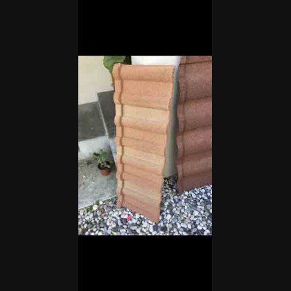 Stack of reddish-brown concrete roof tiles outdoors, leaning against a wall and ground covered in small pebbles.