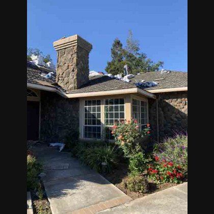 Stone-clad house with bay window, chimney, and roof undergoing repairs, blue sky, plants in front.