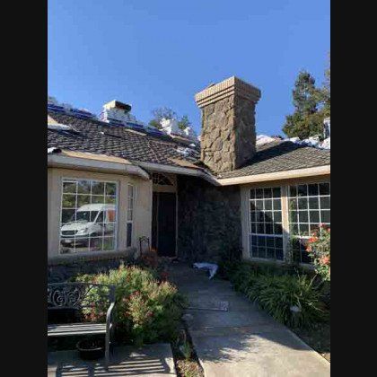 House exterior with stone chimney, windows, and damaged roof.
