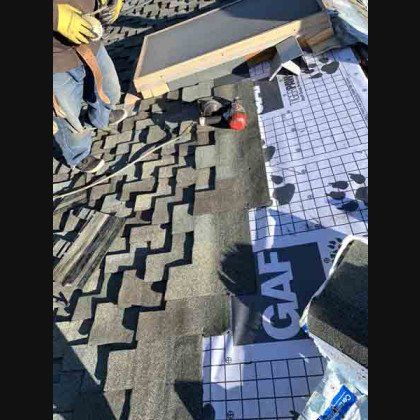 Roofer installing shingles on a roof. They are wearing gloves. Gray shingles and underlayment are visible.