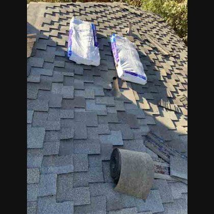 Roof with gray shingles, two bags, and rolled roofing felt, undergoing repair.