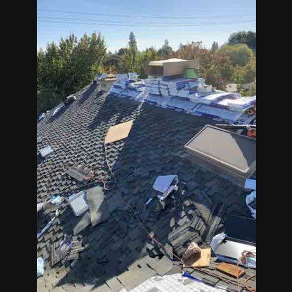 Rooftop with shingles under renovation, piles of new shingles, and equipment. Trees and sky in background.