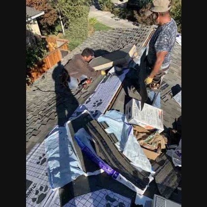 Two men on a rooftop removing shingles and materials. Overcast day.