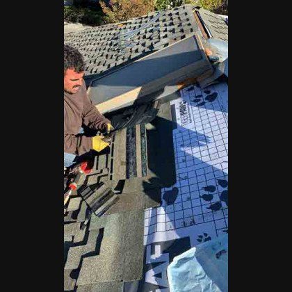 Roofer on a roof, installing shingles near a skylight. Daylight and materials visible.