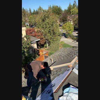 Two roofers working on a rooftop under a sunny sky. Residential neighborhood visible.