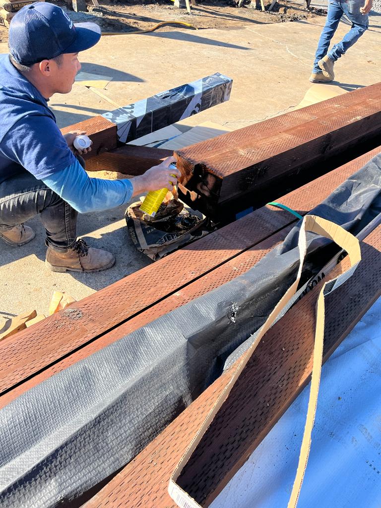 Man inspecting a damaged wooden beam at a construction site. He wears a blue cap and jacket.