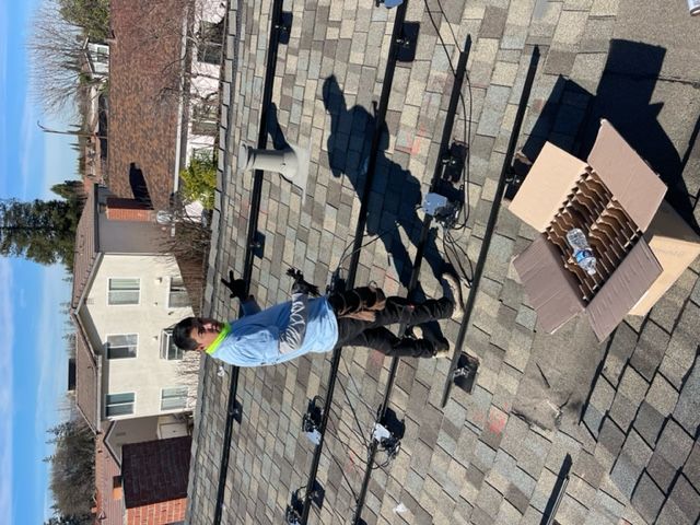 Man on a roof installing solar panels on a sunny day. He's wearing a blue shirt and holding tools.