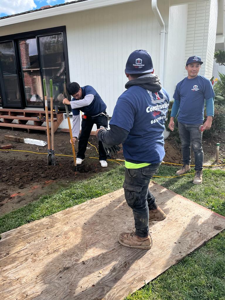 Three construction workers outdoors, one digging, one observing, and one standing on a wooden board.