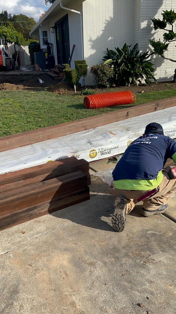 A worker in safety gear kneels, cutting wood outside a house; lawn and construction equipment in the background.