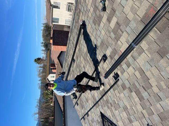 A worker installs solar panel racks on a shingled roof under a bright blue sky.