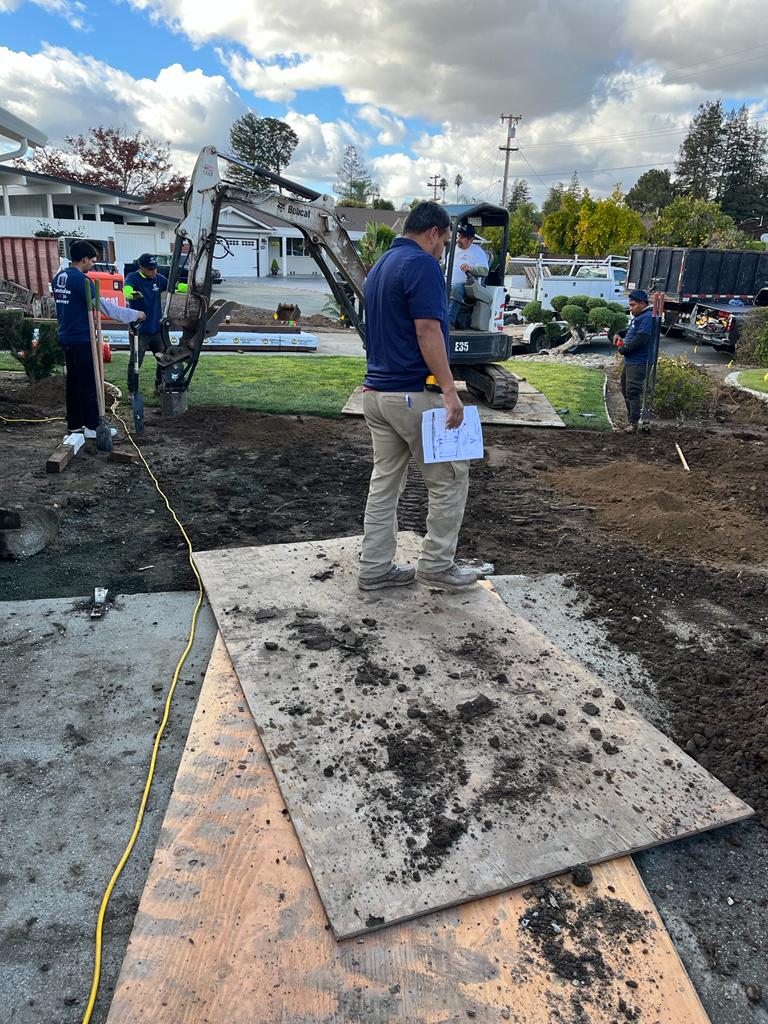 Construction site: workers, excavator, dirt, and plywood in front yard on a cloudy day.