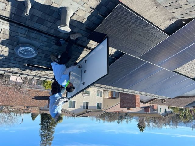 Two workers installing a solar panel on a rooftop. Clear blue sky, houses in the background.