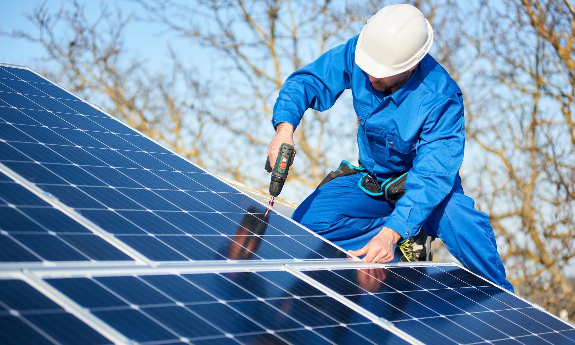 Solar panel installer in blue jumpsuit, using a drill on a rooftop under a blue sky.