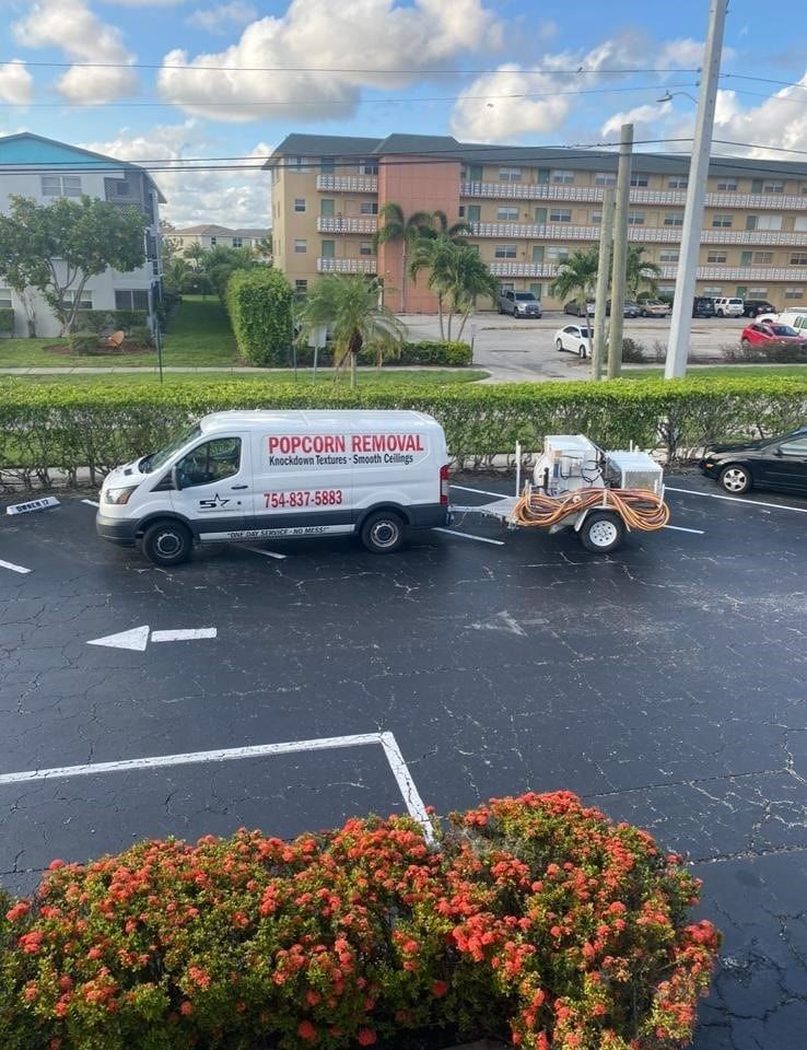 White rental van towing a small equipment trailer on a wet street beside apartment buildings.