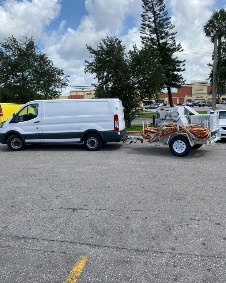 White van parked beside a utility trailer in a parking lot with trees and buildings in the background