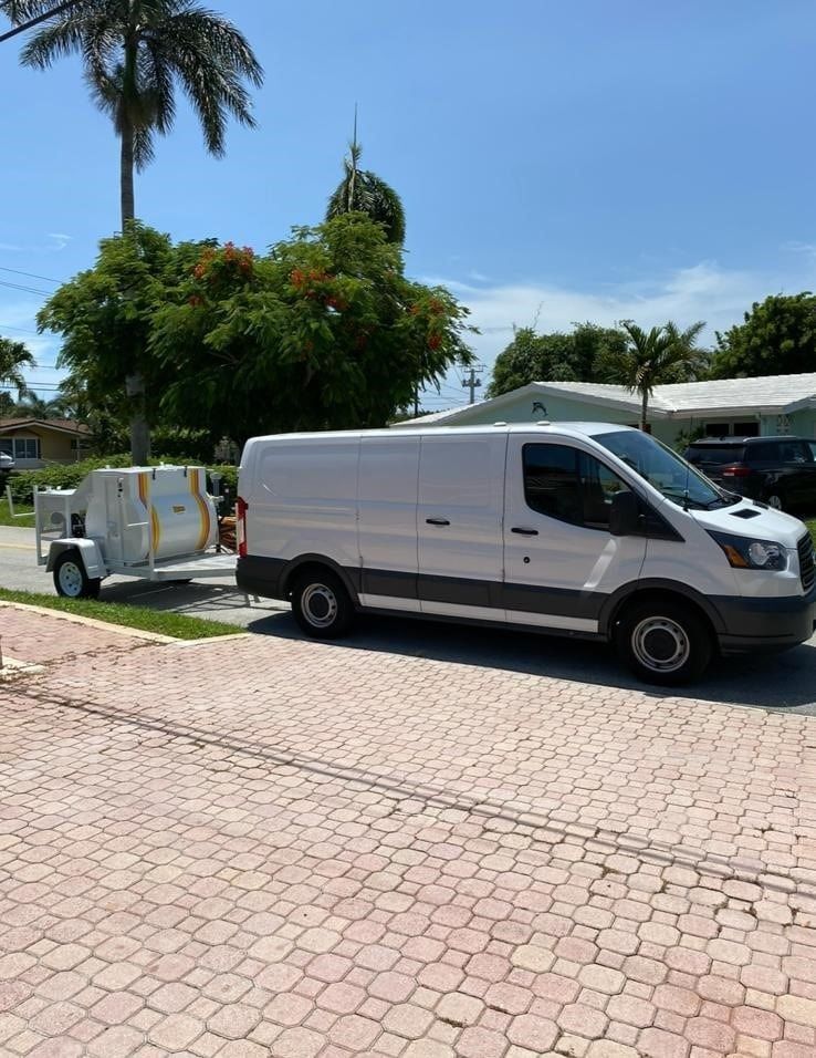White cargo van parked on a sunlit brick driveway beside a small utility trailer and palm trees.