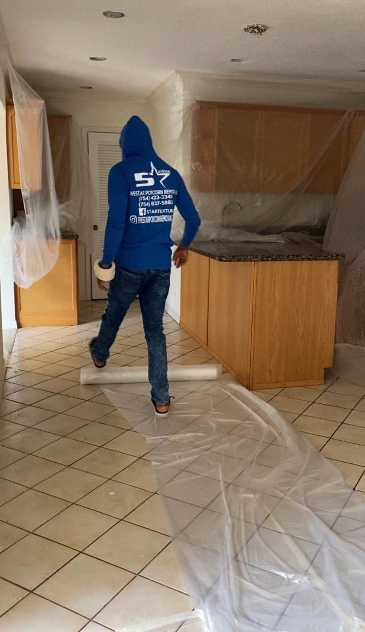 Person in a blue hoodie walking through a kitchen under renovation, with plastic covering floors and cabinets.