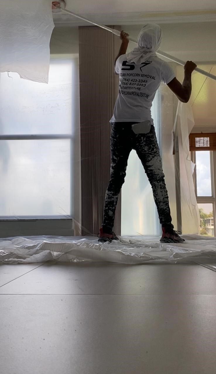 Person painting a wall in a bright room, standing on plastic sheeting with a roller.