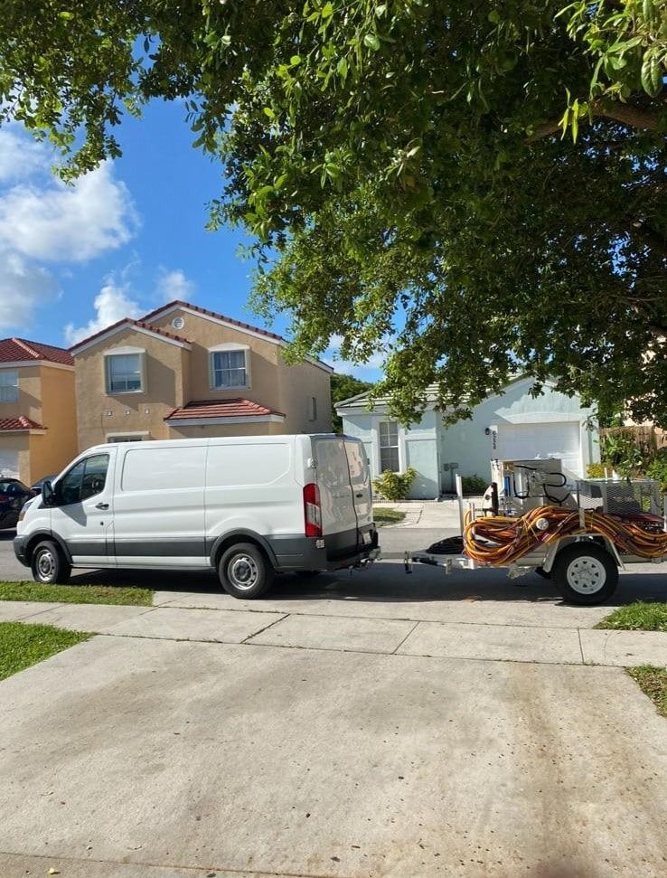 White van towing an orange utility trailer in a suburban driveway under a leafy tree.