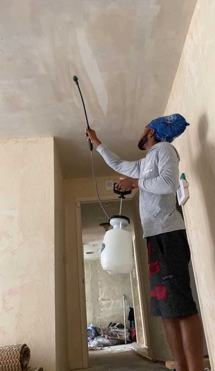 Worker painting a ceiling with a sprayer in a hallway, wearing a blue head covering and white shirt.