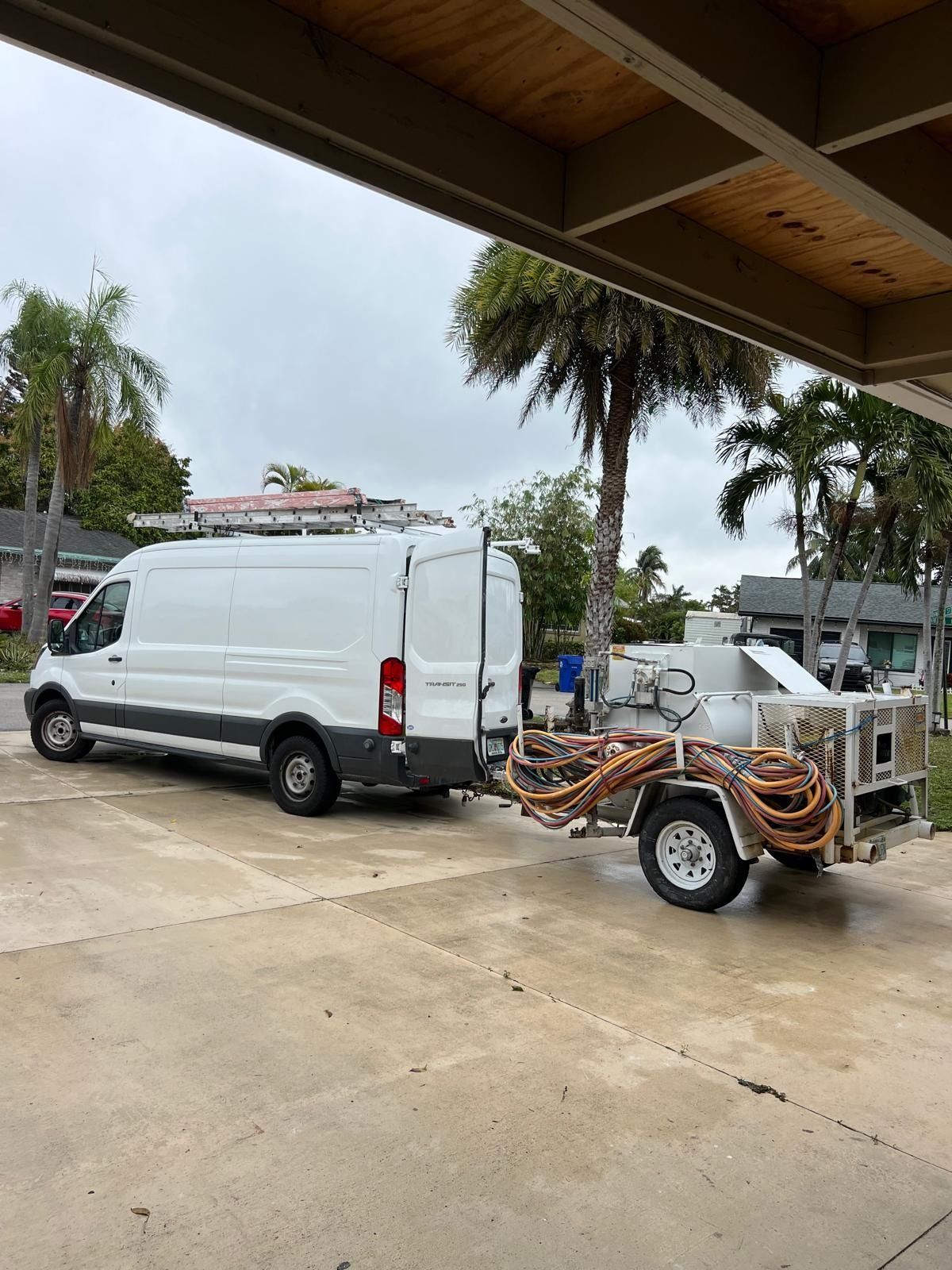 White van and utility trailer parked under a shelter on a wet concrete lot, with palm trees in the background
