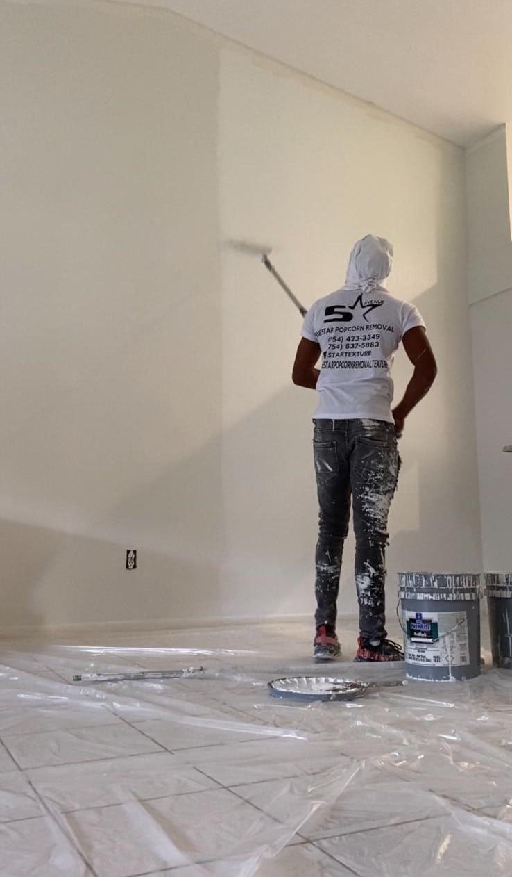 Person painting a white wall in a room covered with plastic drop cloths.