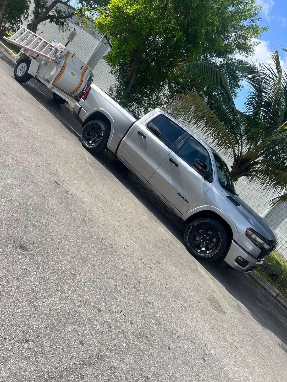 Silver pickup truck parked beside a small utility trailer on a sunny street, with trees in the background