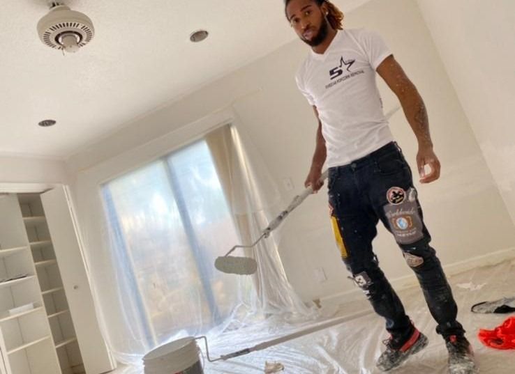 Man painting a bright white room with a roller, floor covered in protective plastic