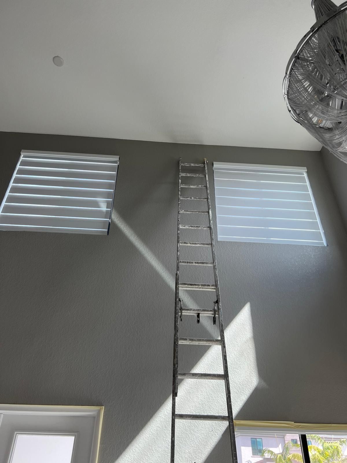 Tall ladder reaching a high wall between two windows, with sunlight casting shadows near a ceiling fan