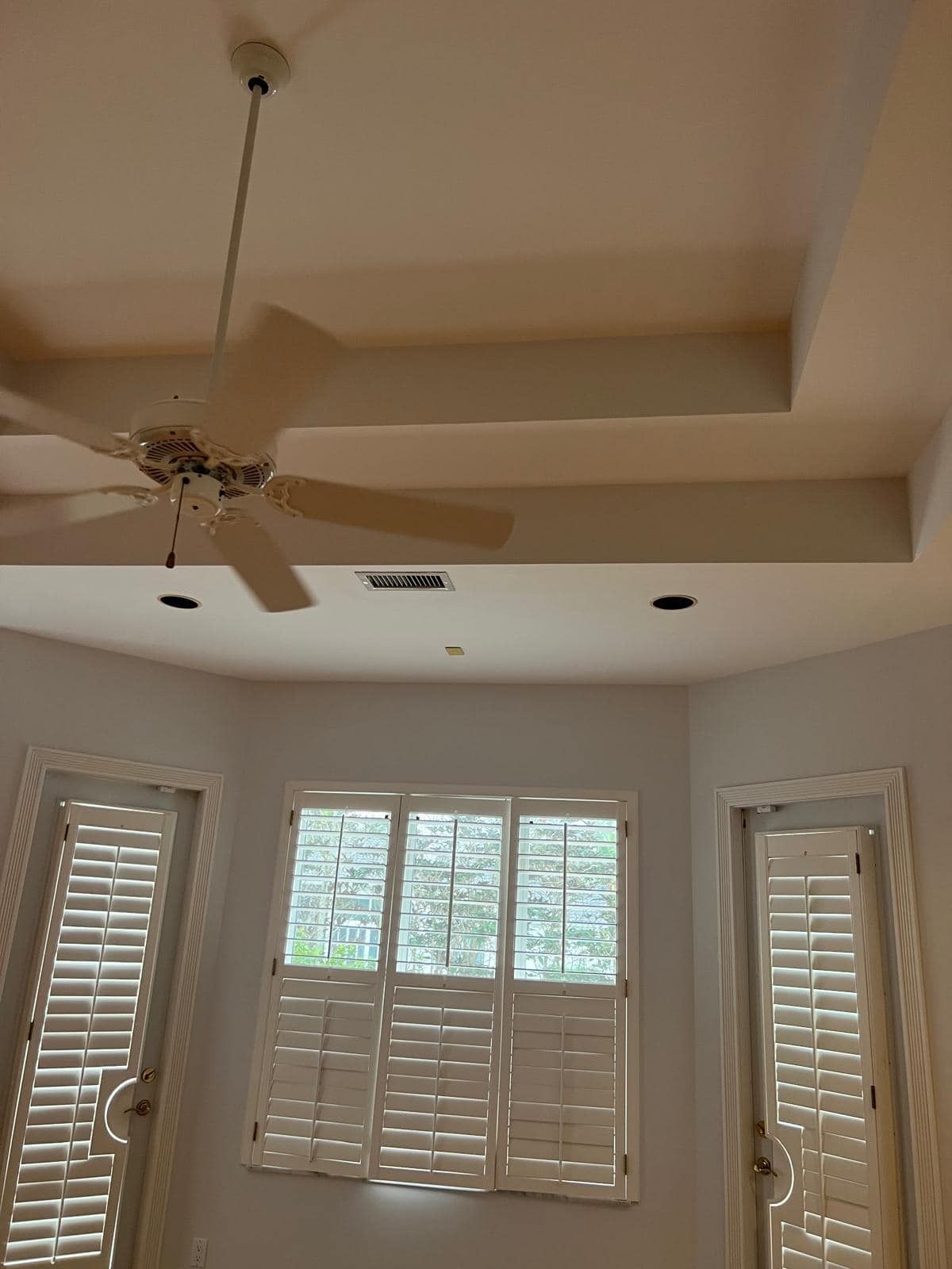 Bright bedroom ceiling with fan and tray molding, above windows with white shutters and blinds.