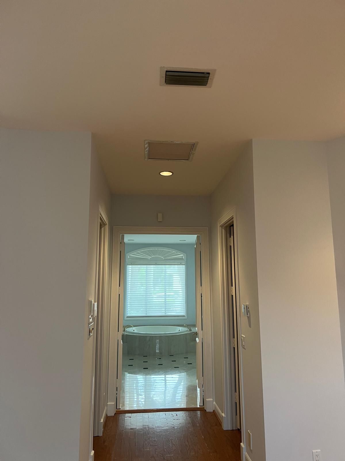 Narrow hallway with white walls and wood floor, leading to a bright room at the end.