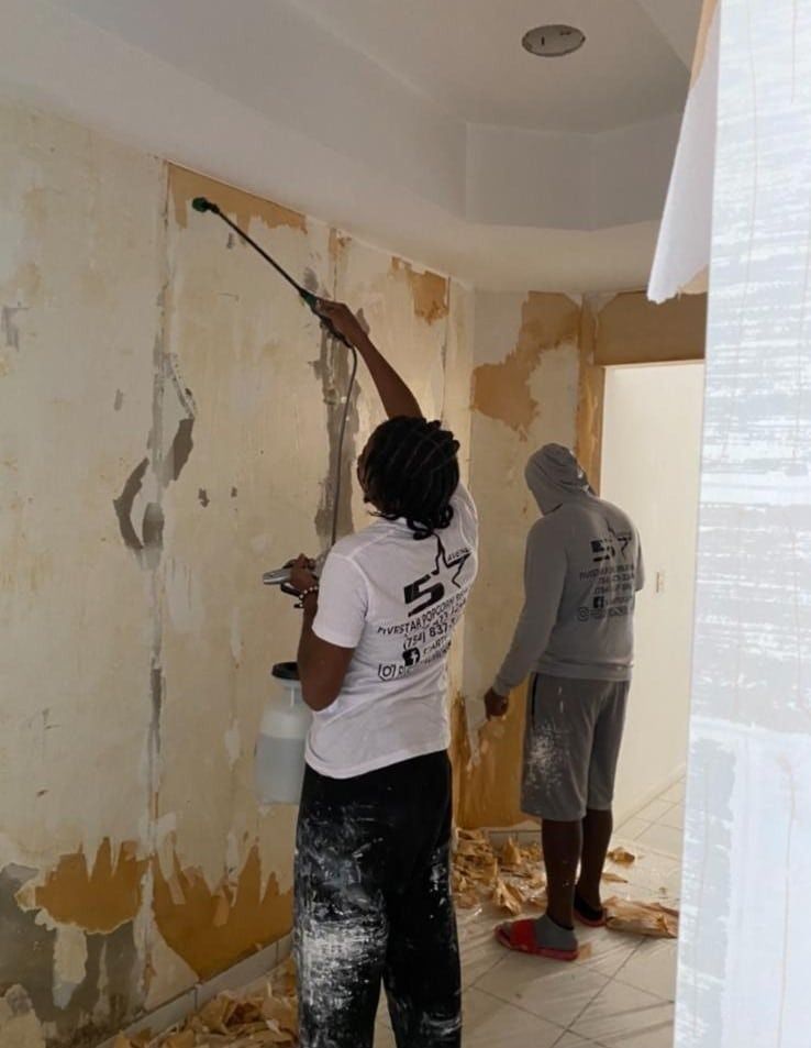 Two people painting a damaged wall and ceiling in a room with peeling paint and a drop cloth on the floor.