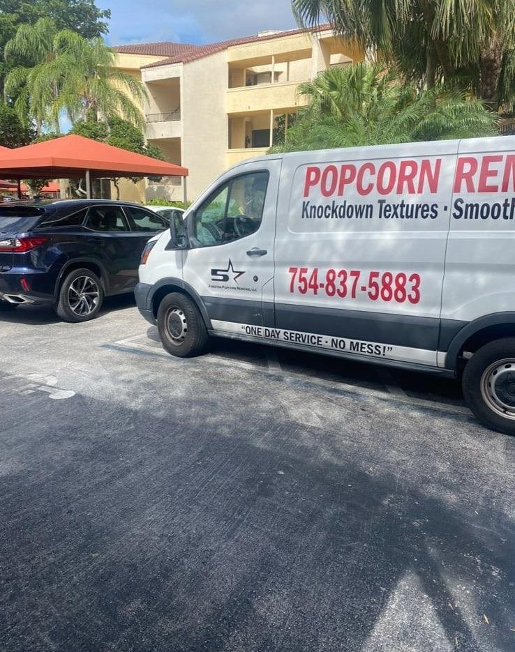 White popcorn rental van with red lettering parked beside a black SUV near a hotel and palm trees.