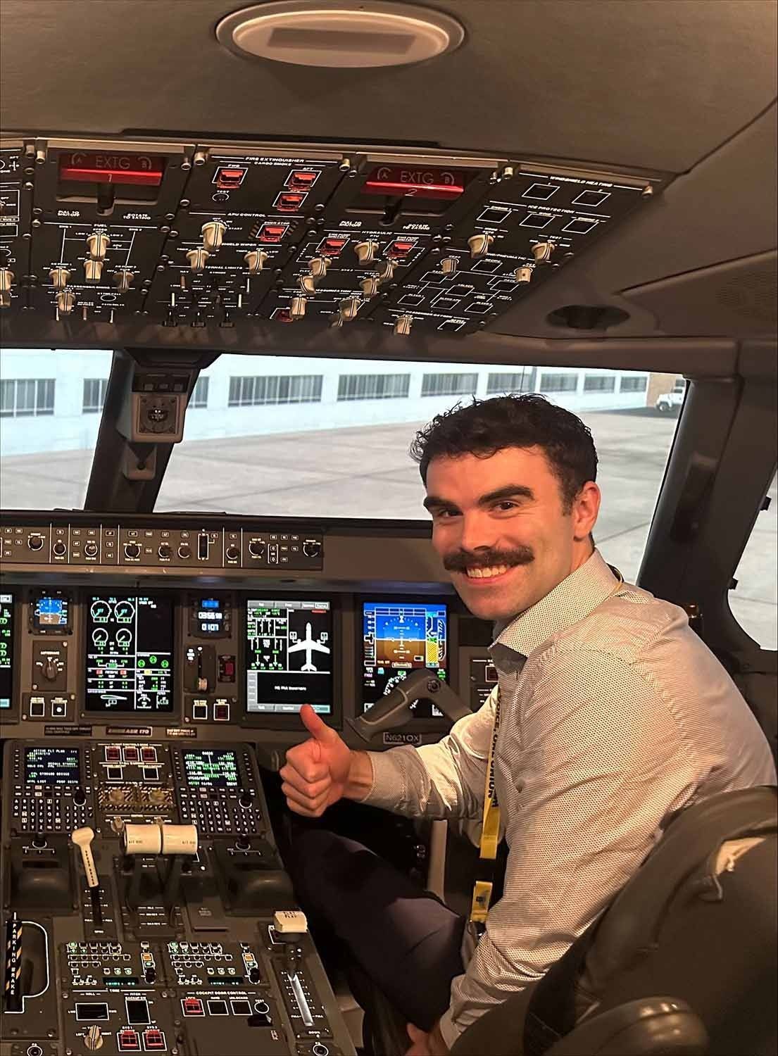 A man with a mustache is giving a thumbs up in the cockpit of an airplane.