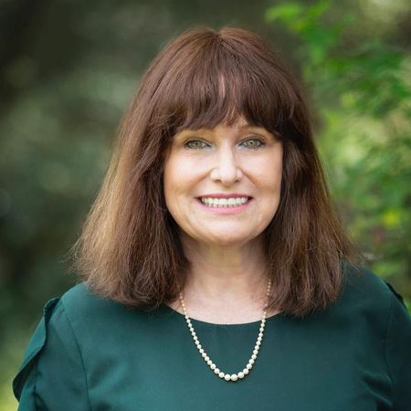 Smiling woman with brown hair in a green top and pearl necklace outdoors, against a blurred green background