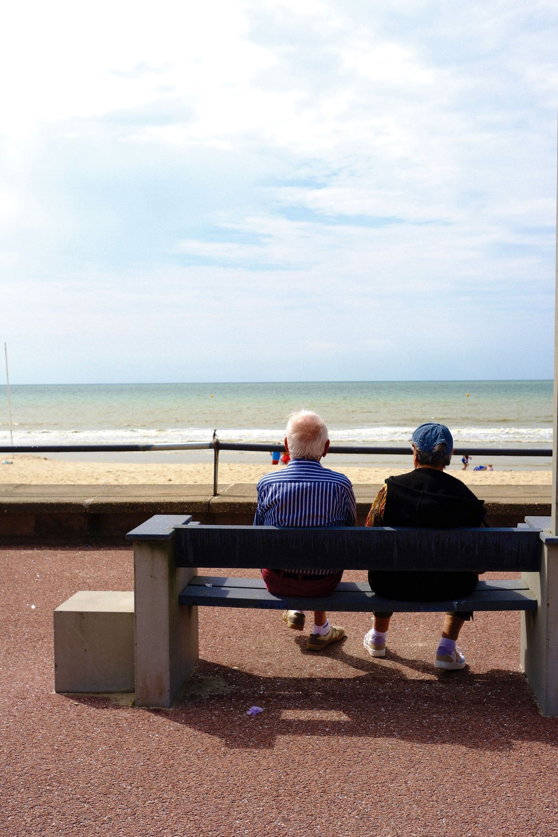 Two people sitting on a bench facing the ocean under a bright sky.