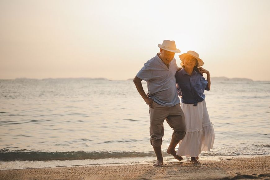 Couple walking barefoot on the beach at sunset, wearing hats, with calm ocean waves behind them