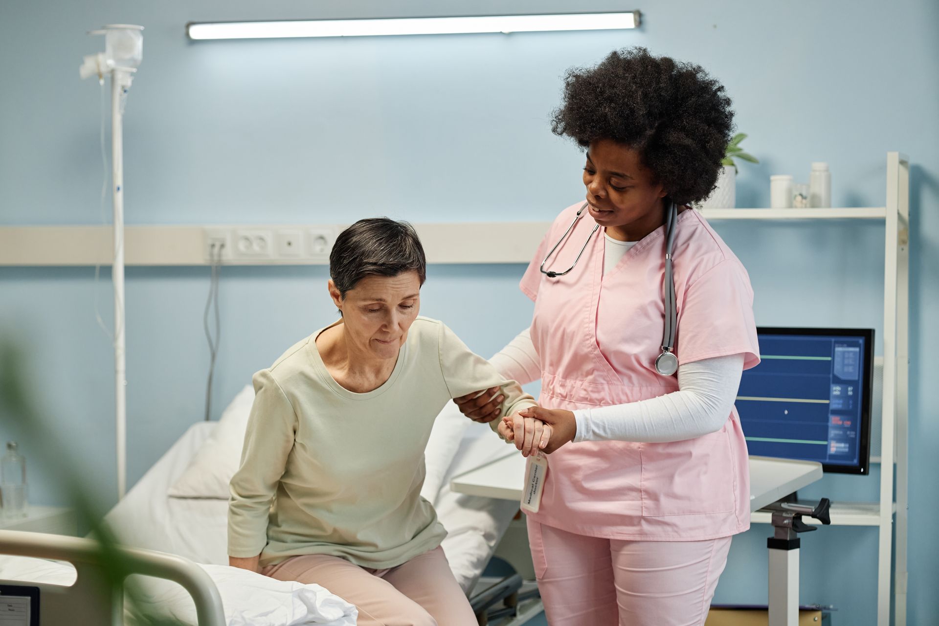 Nurse in pink scrubs assists a patient seated on a hospital bed in a clinic room.