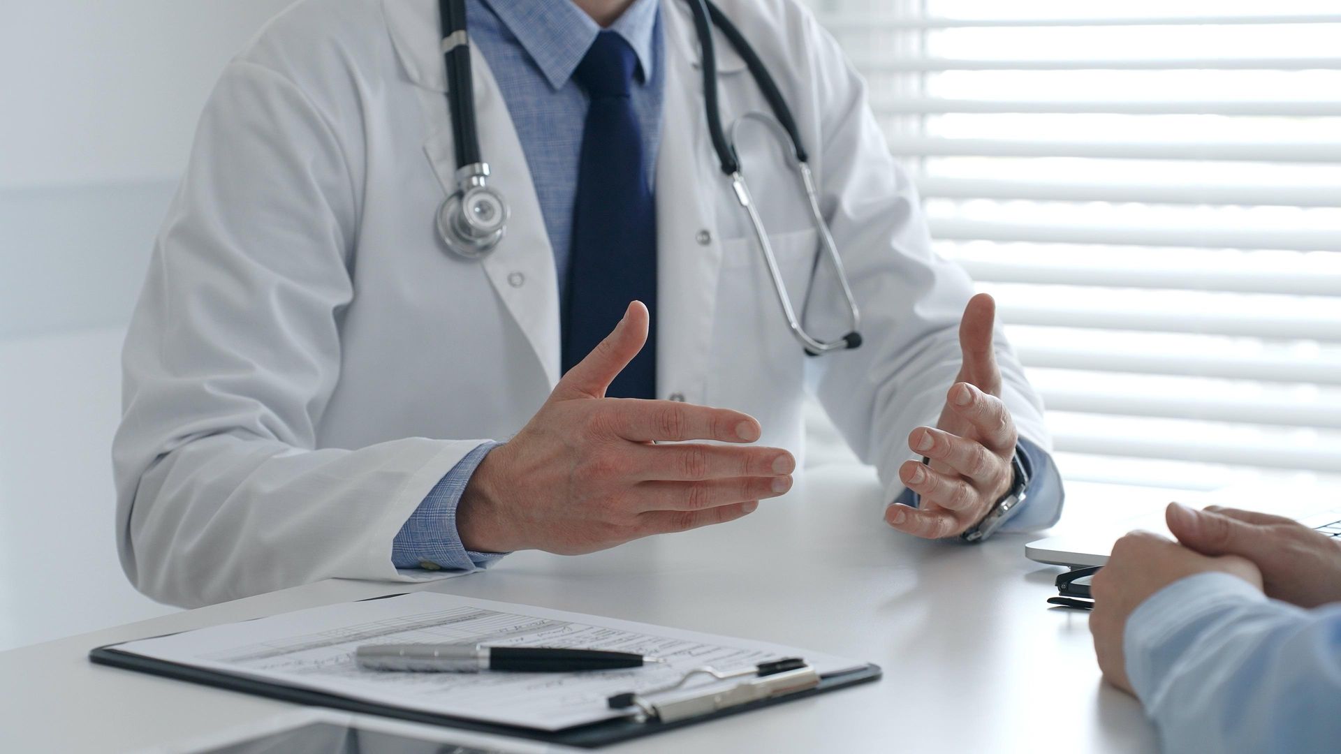 Doctor in white coat and stethoscope speaking with a patient across a desk in a bright office