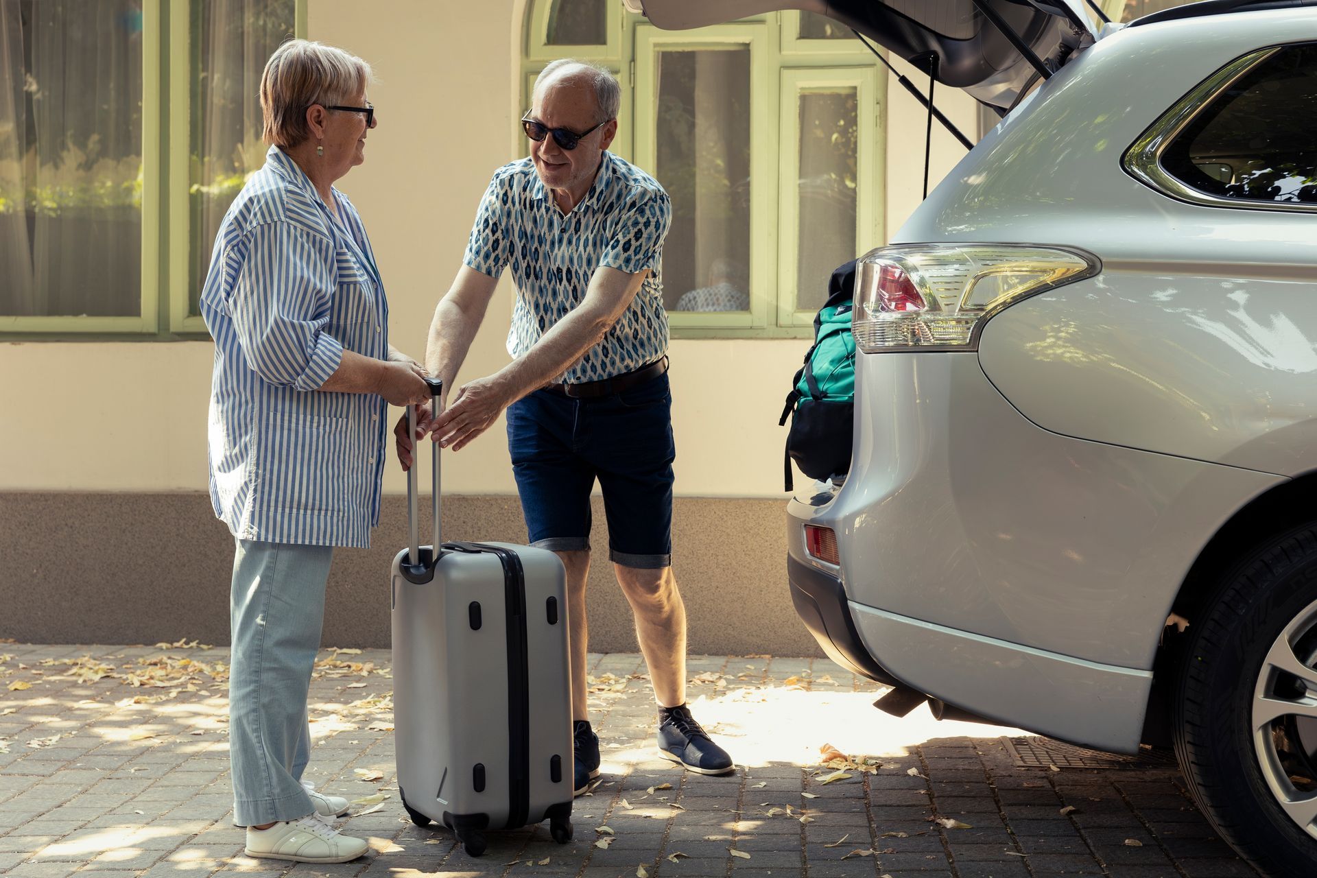 Two people unloading a silver SUV, standing by a suitcase on a driveway.