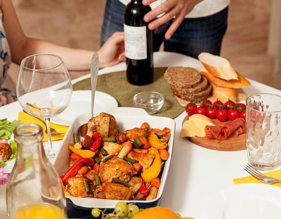 Shared meal table with roasted vegetables, bread, grapes, wine, and hands pouring a bottle