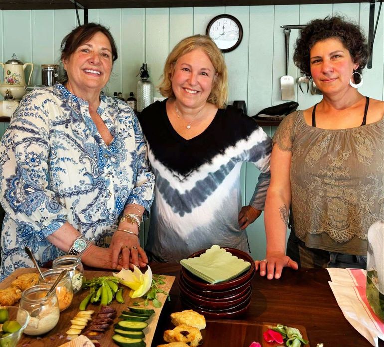 Three women smiling behind a table of food in a kitchen