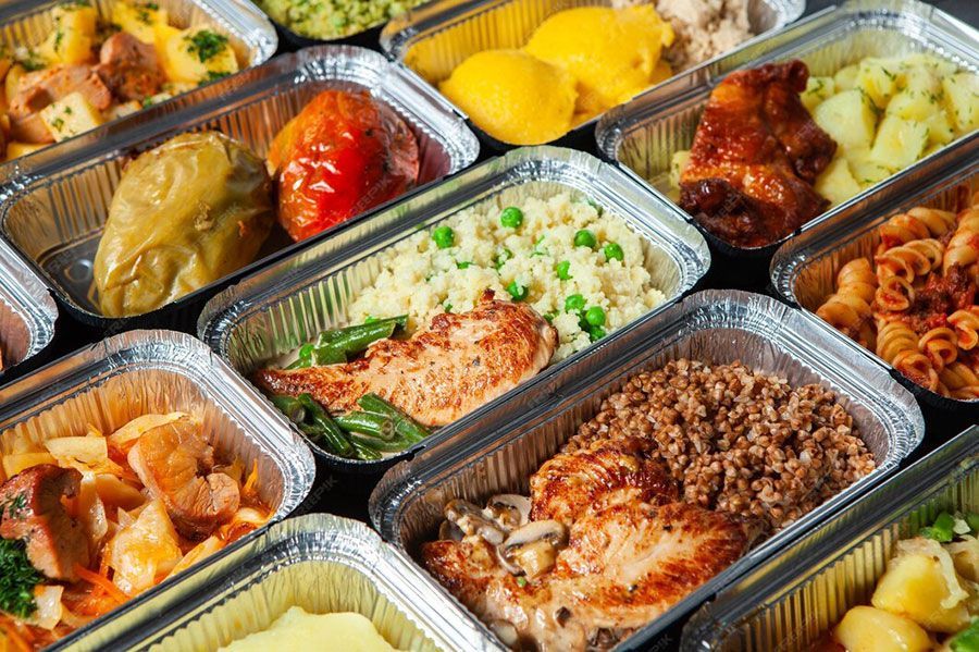 Assorted foil takeout trays with prepared meals, including pasta, chicken, rice, and vegetables, arranged in rows