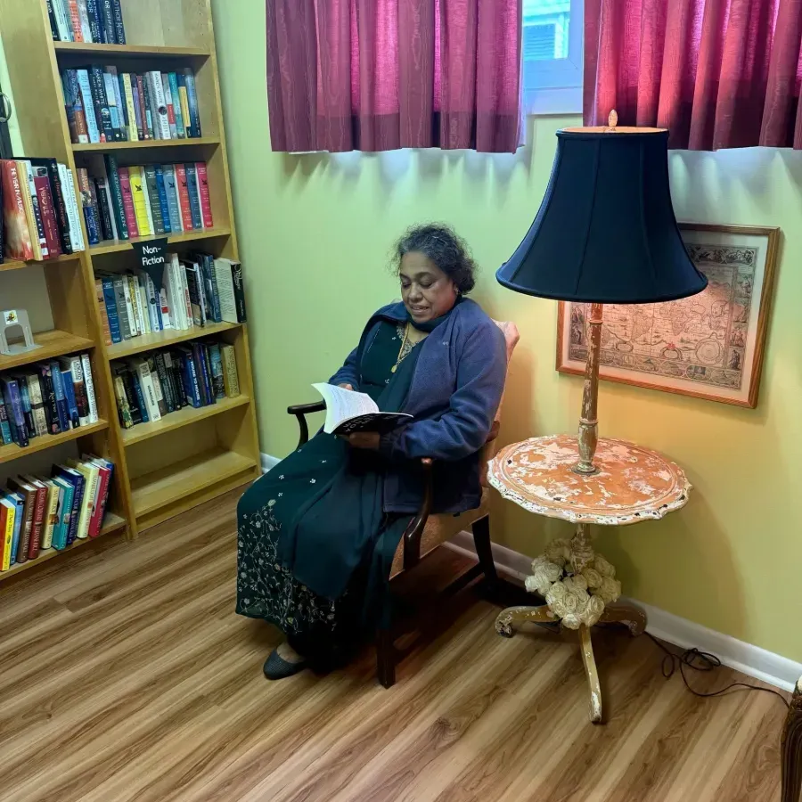 Person reading in a cozy room beside a bookshelf, lamp, and framed wall art.