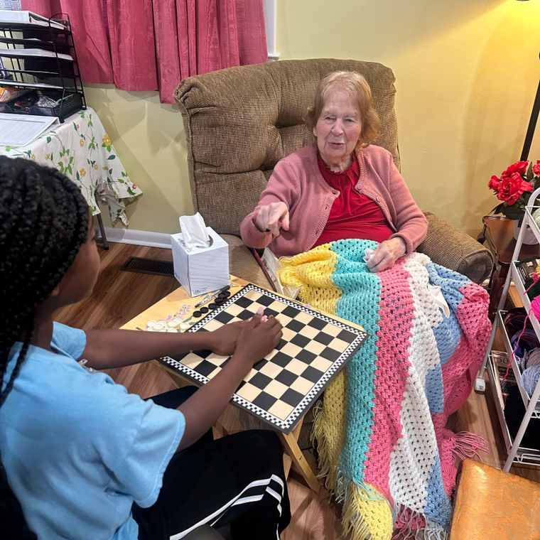 Two people play chess at a wooden table in a cozy room with floral wallpaper.