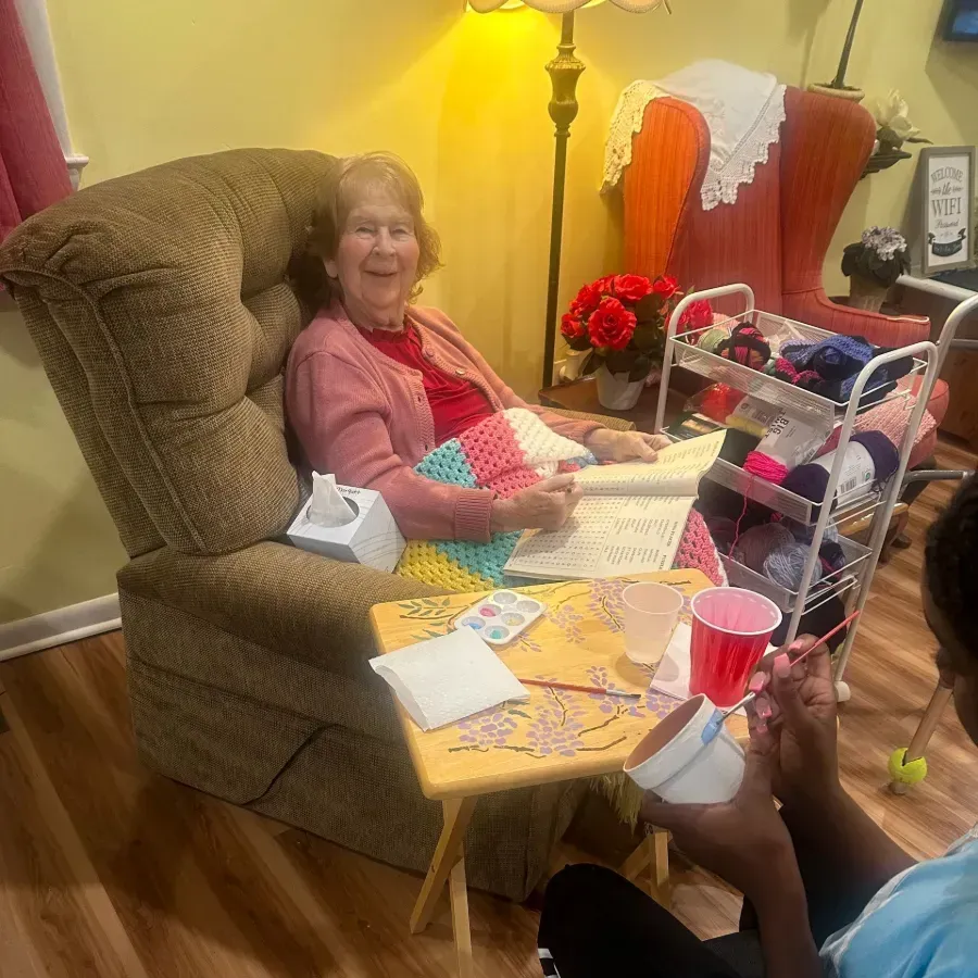 Woman smiling in a recliner beside a craft table with greeting cards, lamp, and colorful decorations