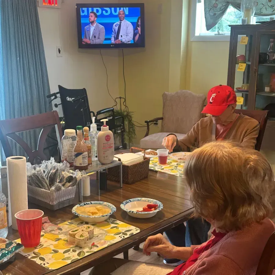 People seated at a dining table with food and drinks in a living room, watching TV.